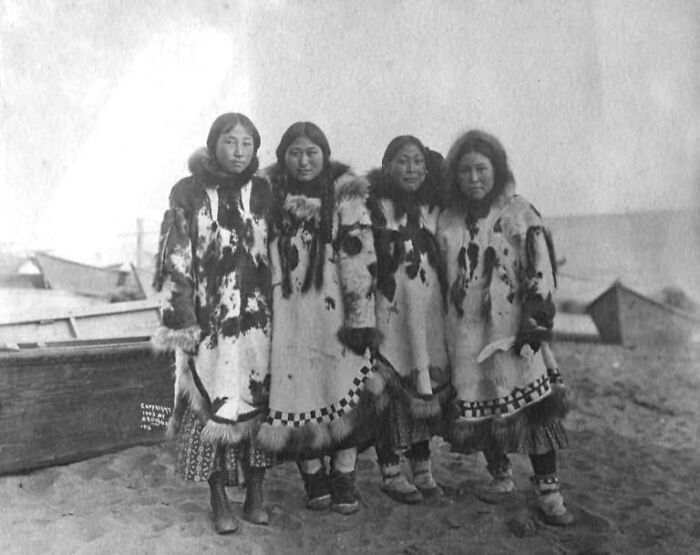 Historic photo of four Inuit women wearing traditional fur clothing standing together near boats on a beach, showcasing Inuit spirit.
