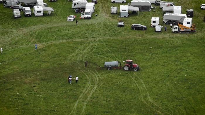 Aerial view of a farm vehicle spraying manure near camper vans and people on a grassy field. Aerial view of a farm vehicle spraying manure near camper vans and people on a grassy field.