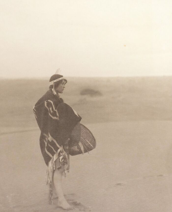 Native tribes member in traditional clothing standing on a sandy landscape, captured in an early 1900s photograph.