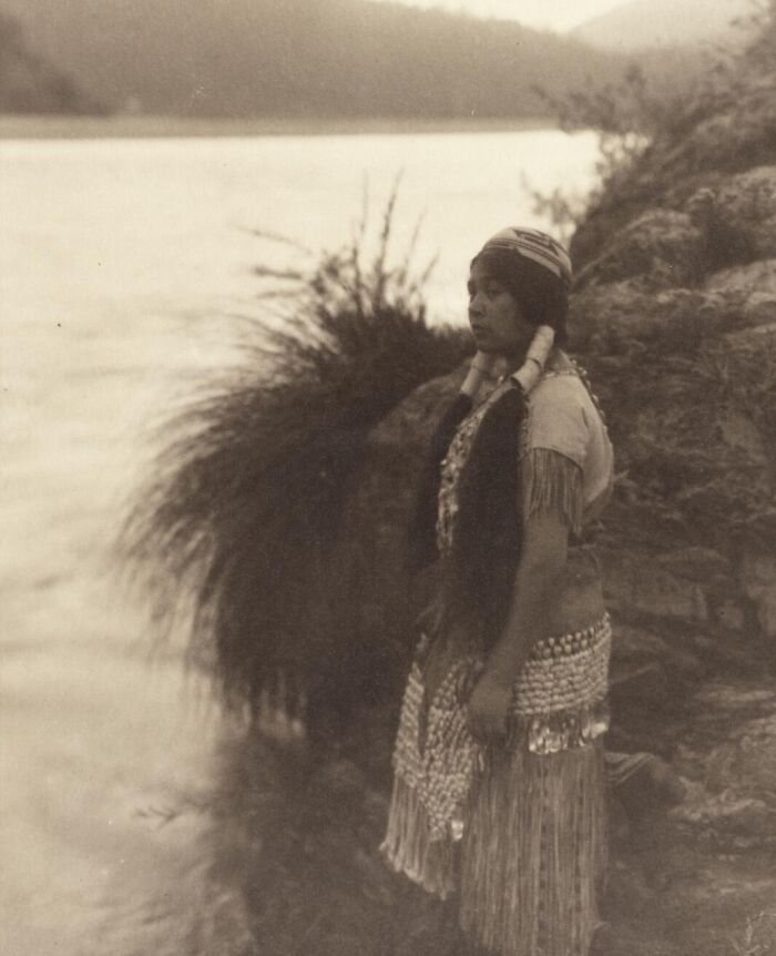 Native tribeswoman standing by a river in traditional dress, captured in a vintage 1900s photograph showing tribal strength.