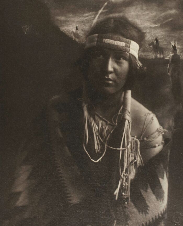Native tribes member in traditional attire with headband and beaded accessories captured in a historic 1900s photograph.