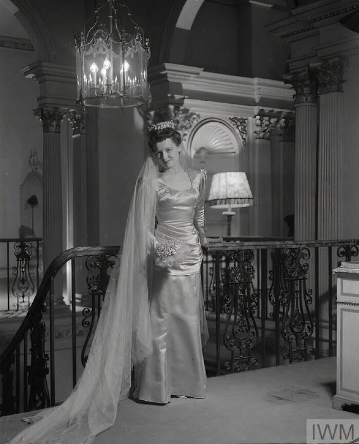 British woman in elegant satin wedding dress and long veil showcasing style during WWII restrictions indoors by staircase.