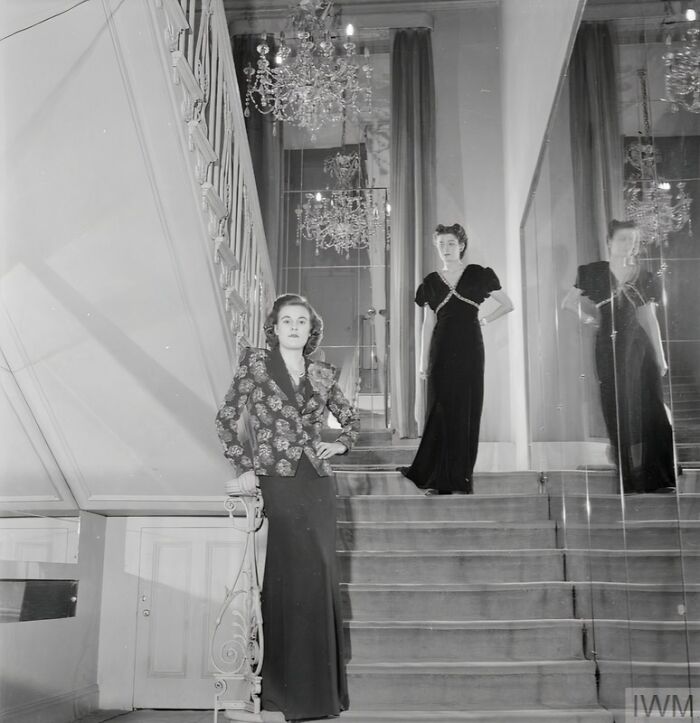 Two British women in elegant dresses posing on a staircase, showcasing style during WWII restrictions.