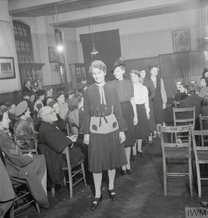 British women showcasing fashion and style during WWII restrictions in a community hall with seated audience watching.