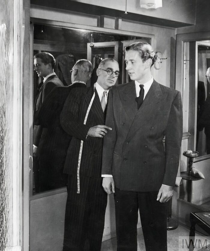 Tailor measuring a man for a suit in a fitting room, illustrating British women's style during WWII restrictions.