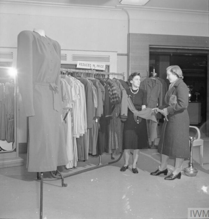 Two British women examining clothing in a store, showcasing style during WWII restrictions and fashion challenges.