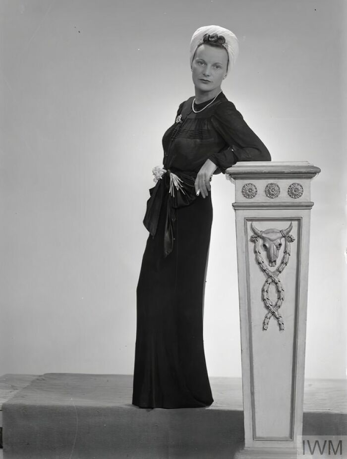 British woman in elegant WWII-era fashion poses beside a decorated pillar, showcasing style during wartime restrictions.