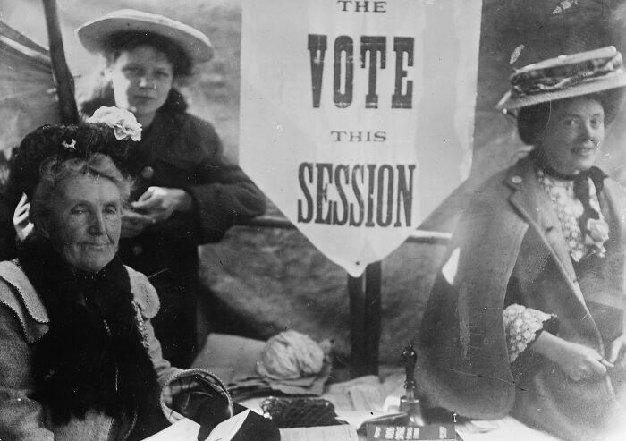 Women in the 20th century suffrage era campaigning for voting rights, dressed in period clothing at a voting session booth.