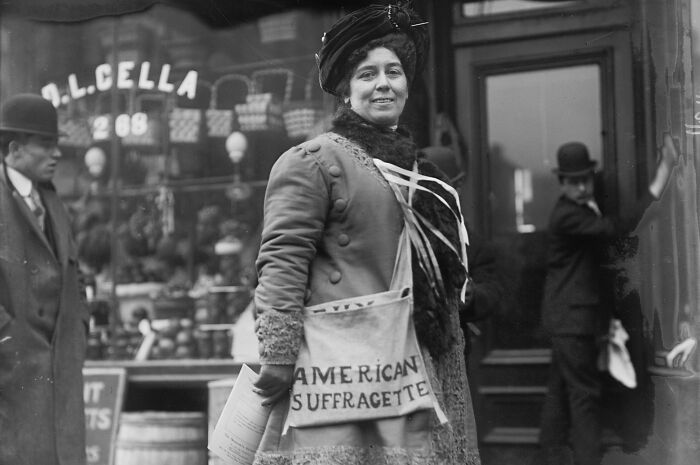 Woman wearing American suffragette sash during the 20th century suffrage era, standing on a city street with others nearby.