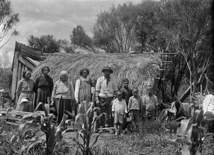 Portrait of a Māori family standing in front of a traditional thatched house, showcasing Māori life a century ago.