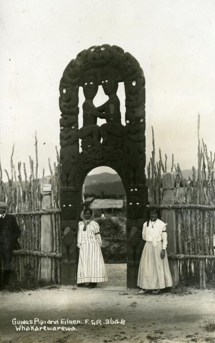 Two Māori women standing under a large carved wooden archway showcasing Māori life and culture a century ago.