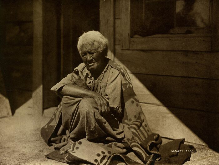 Elderly Māori man sitting wrapped in a patterned cloak, smoking a pipe in a sepia-toned historic portrait.