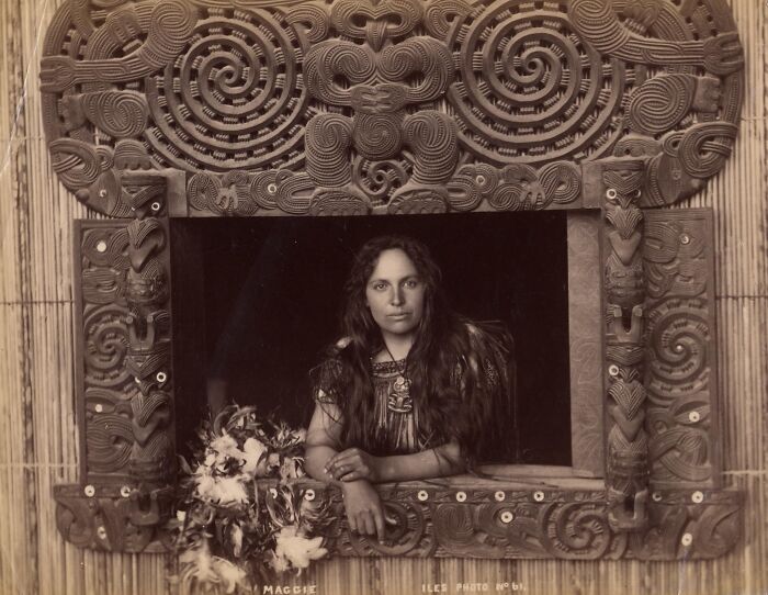 Māori woman in traditional attire framed by intricate carved wooden panels representing Māori life a century ago.
