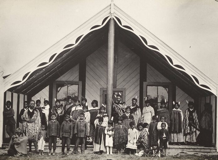Group portrait of Māori men, women, and children in traditional clothing in front of a meeting house from a century ago.