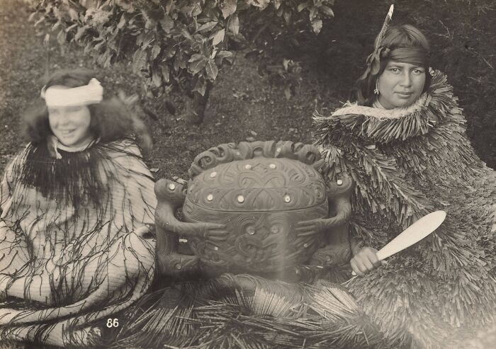 Two Māori individuals in traditional attire beside a carved wooden drum, capturing Māori life a century ago.