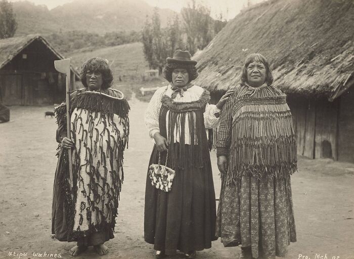 Three Māori women in traditional clothing standing outdoors near thatched huts in a powerful portrait of Māori life a century ago.