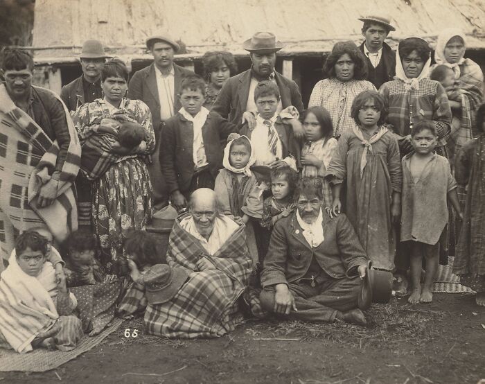 Group portrait of Māori life showing multiple generations in traditional and early 20th-century clothing outdoors near a thatched building