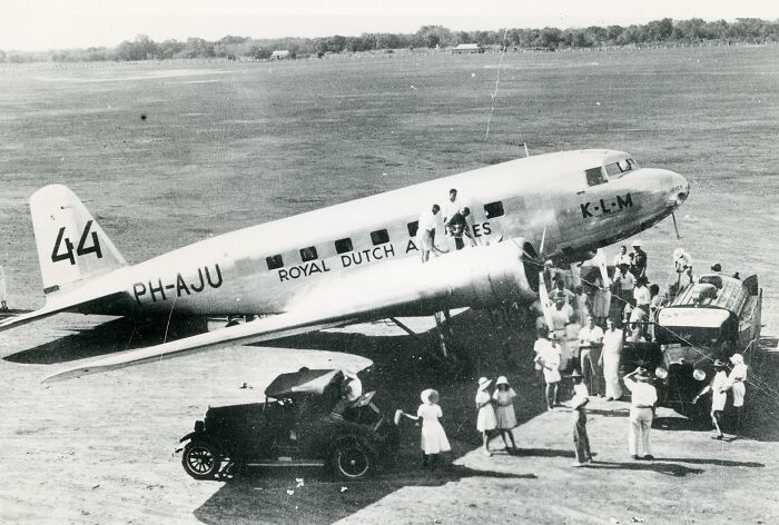 Vintage black and white photo of a Royal Dutch Airlines plane with passengers and classic cars, showing glamorous flying era.