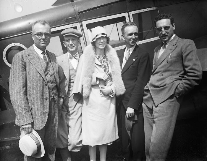 Group of stylishly dressed men and a woman posing by a vintage airplane, showcasing flying glamour from a bygone era.