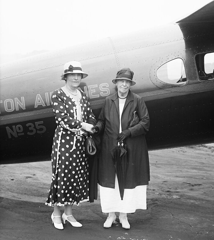 Two elegantly dressed women standing by a vintage airplane, capturing the glamour of early flying experiences.
