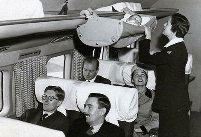 Flight attendant placing a baby in an overhead bassinet during a glamorous vintage flight experience.