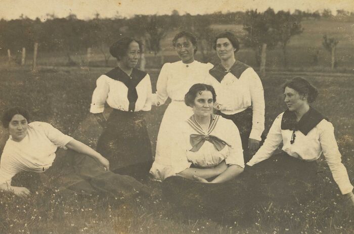 Group of young women at Native American boarding school in early 1900s, wearing period uniforms and posing outdoors on grass.