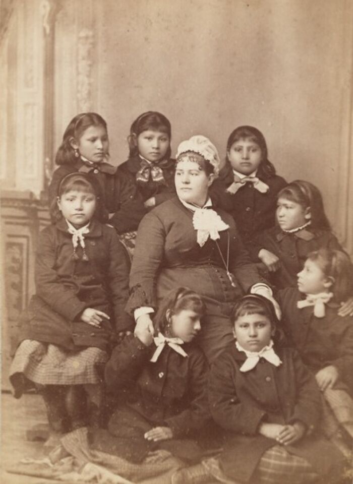 Native American boarding school children from the 1910s, dressed in uniform with a teacher in a formal studio portrait.