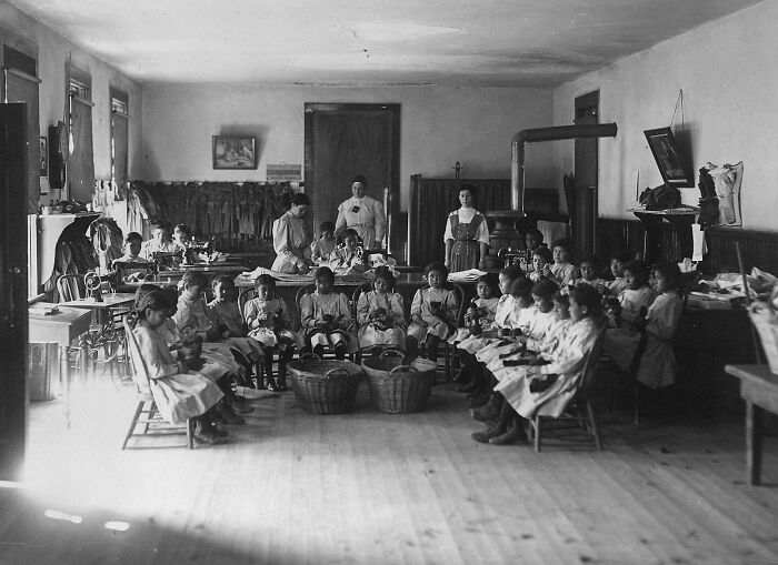Native American boarding school classroom in the 1910s with children seated in a circle, engaged in an activity.
