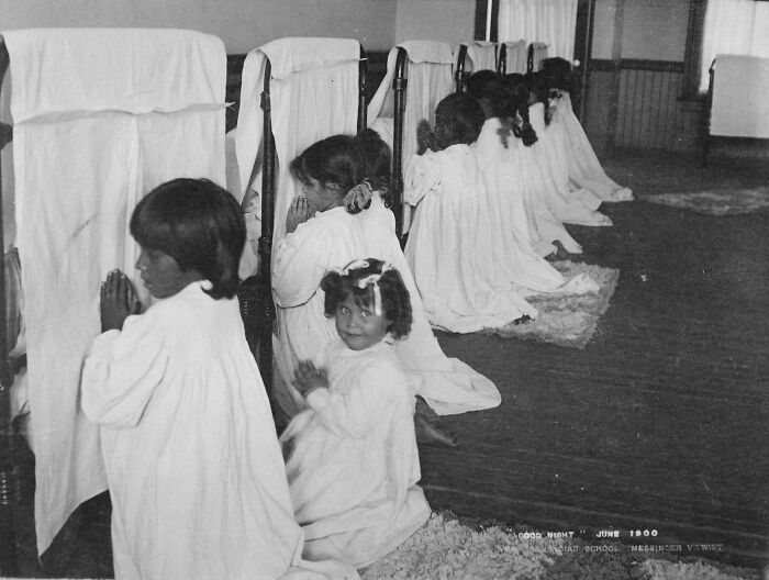 Native American children in white gowns kneeling in prayer at a boarding school in the early 1900s hallway.