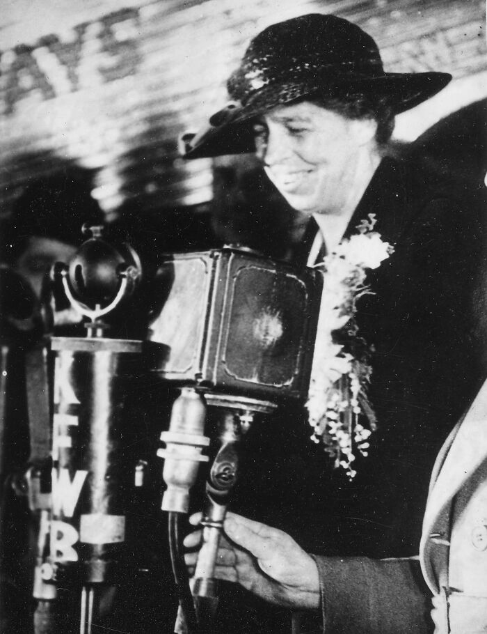 Black and white photo of a smiling LA woman in 1962 wearing a hat and speaking into a vintage microphone.