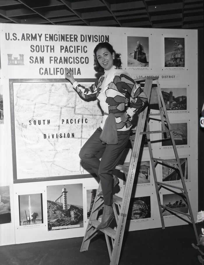 LA woman in 1962 standing on ladder by South Pacific Division map, smiling and posing confidently indoors.
