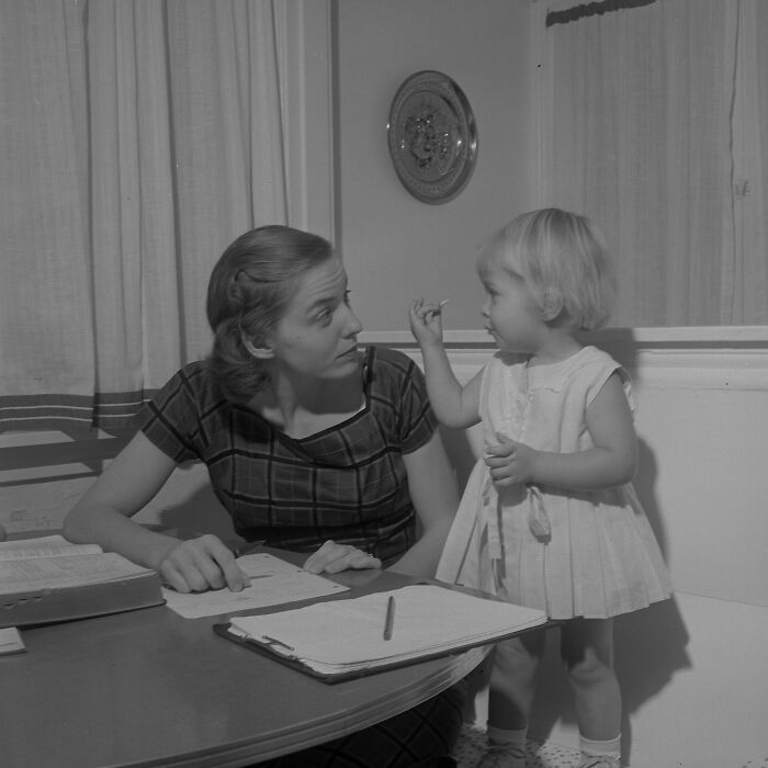LA woman in 1962 sitting at a table with paperwork while interacting with a young child in a home setting.