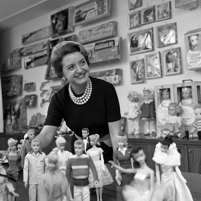 Smiling LA woman in 1962 arranging dolls in a room filled with vintage toys and doll packaging on the walls.