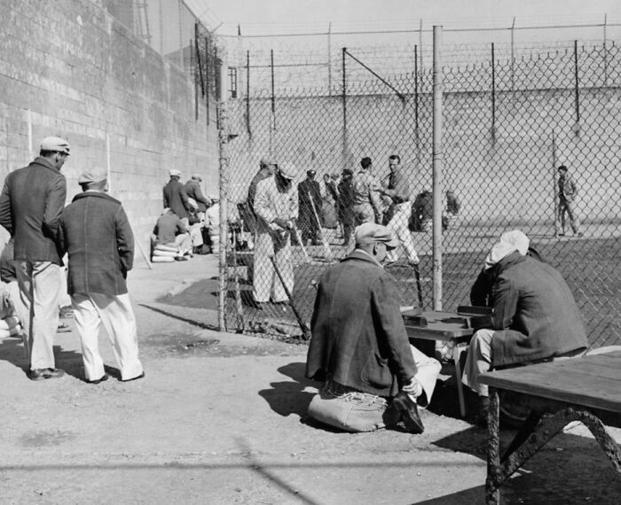 Prisoners in worn clothing inside Alcatraz yard, some playing games and others standing near the chain-link fence.