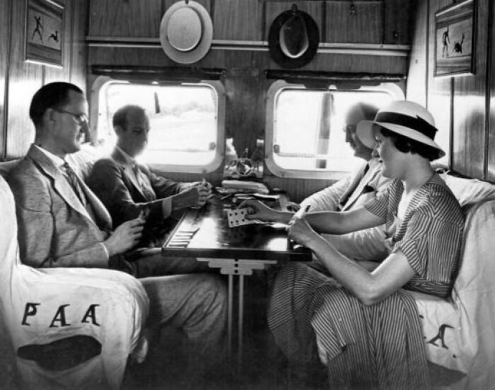 Passengers dressed in vintage attire playing cards aboard a luxurious airplane interior, showcasing flying glamour in the past.
