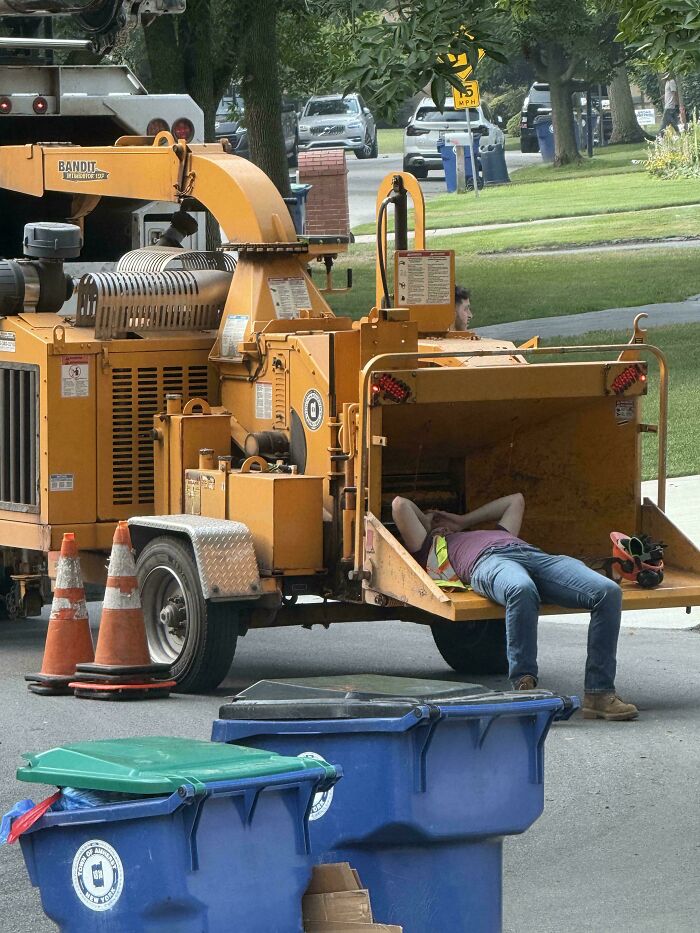 Worker dangerously lying inside wood chipper ignoring safety protocol near street cones and trash bins.