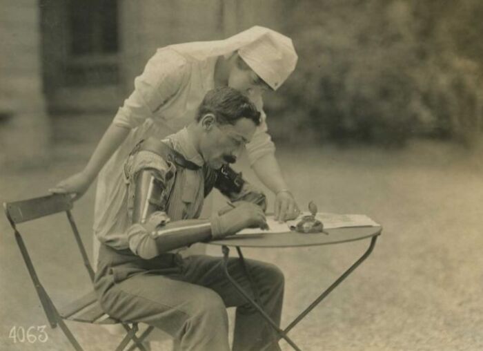 WWI daily life showing a soldier with a prosthetic arm writing at a table, assisted by a nurse outdoors.