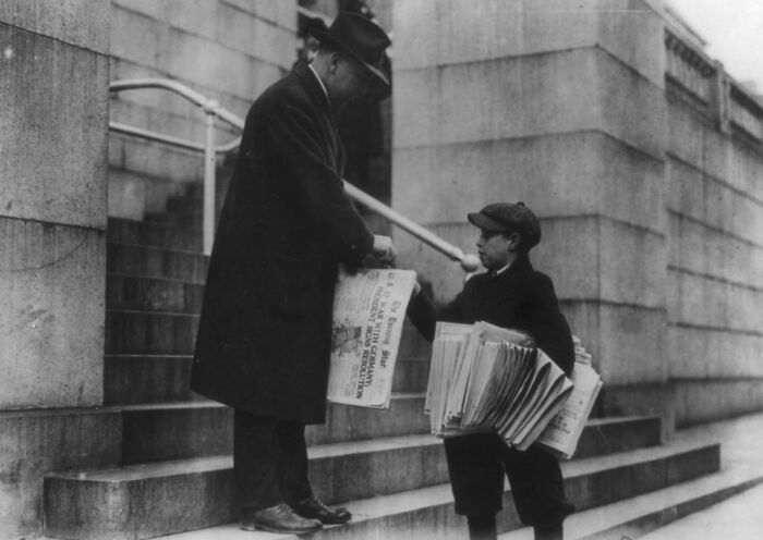 Boy selling newspapers on city steps to a man in coat and hat, showing daily life beyond WWI trenches in urban setting.