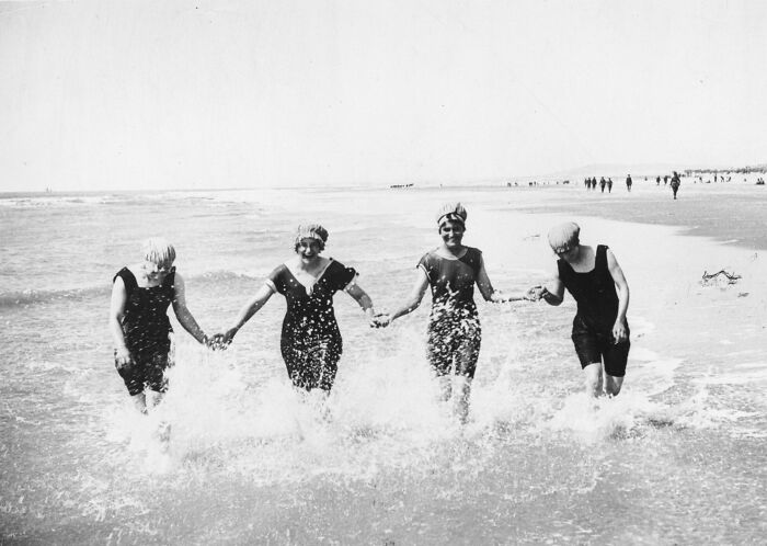 Four women in vintage swimwear holding hands and splashing water on a beach, showing daily life beyond WWI trenches.