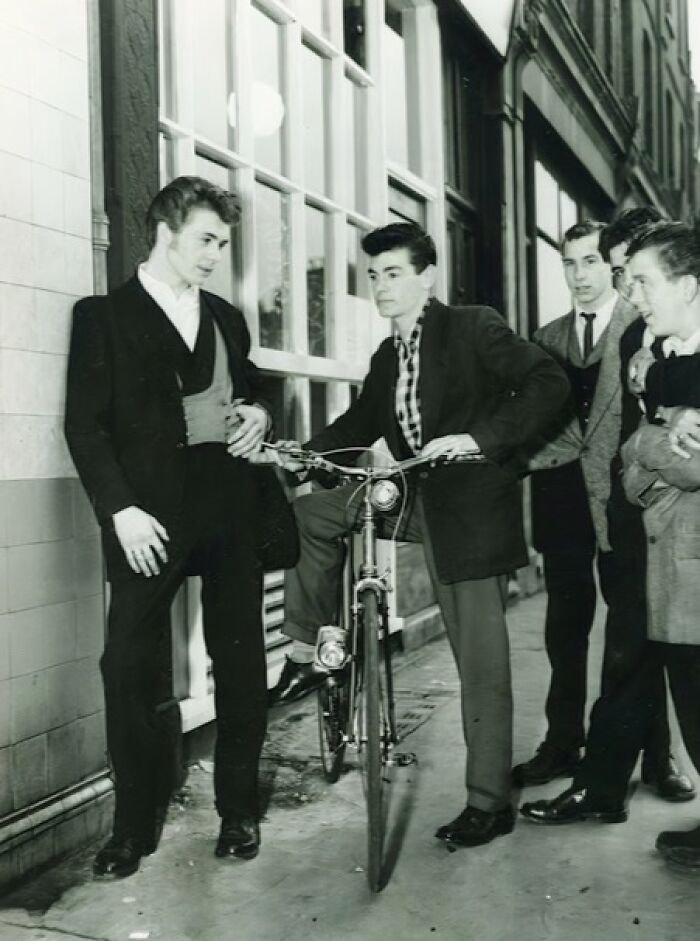 Group of young men in vintage Teddy Boy style clothing, one posing on a bicycle in a narrow alleyway.