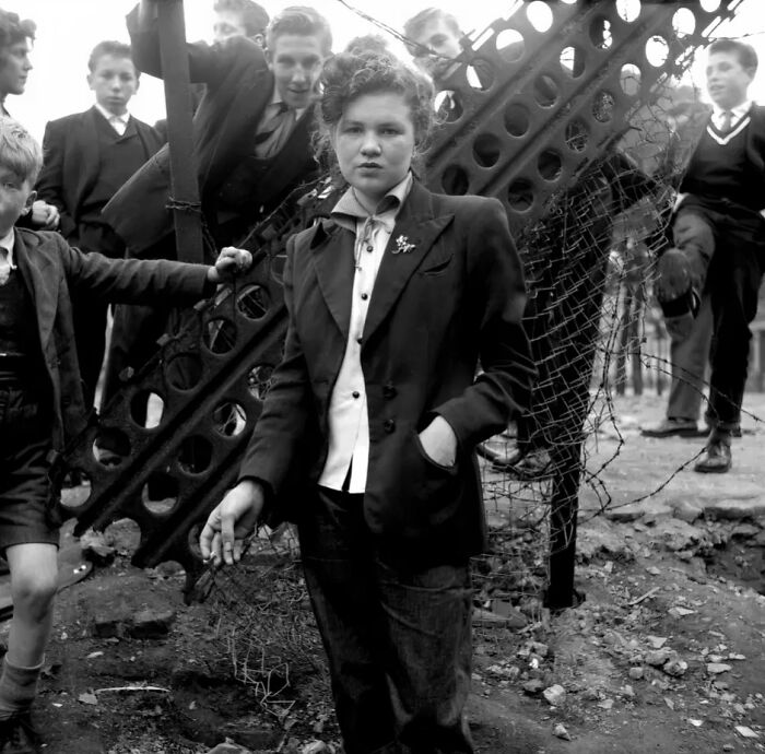 Teenagers dressed in classic 20th century Teddy Boy era style pose outdoors near broken metal and wire fence.