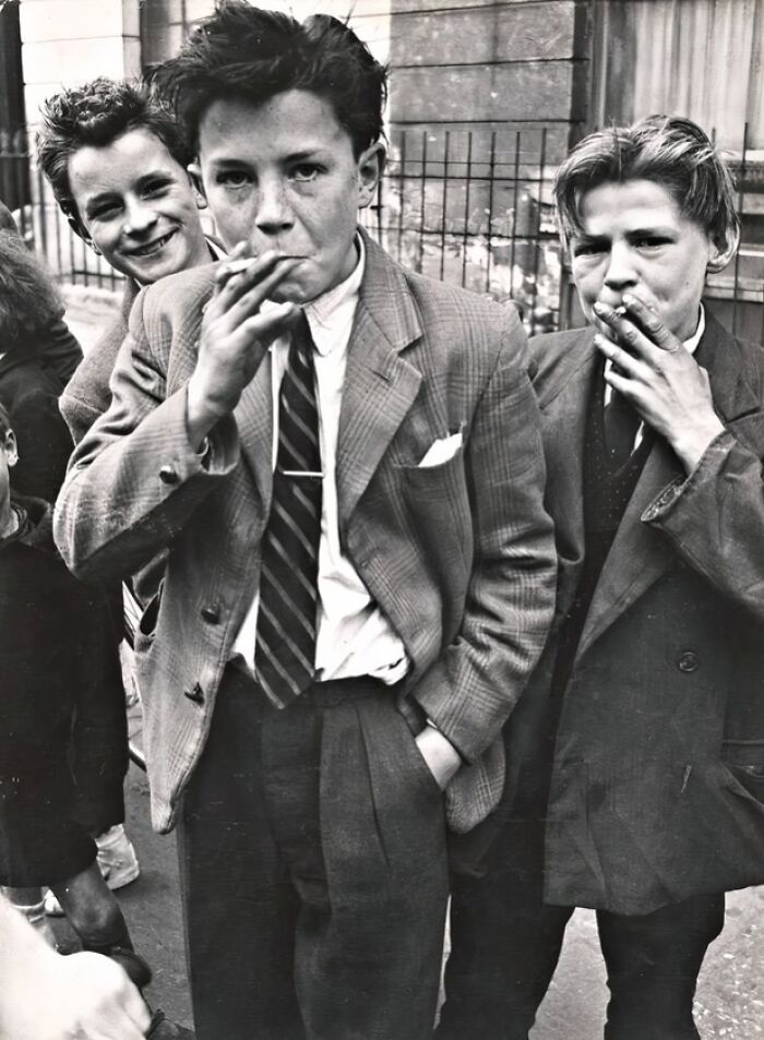 Three teenage Teddy Boys in vintage suits smoking cigarettes on a street during the 20th century Teddy Boy era.