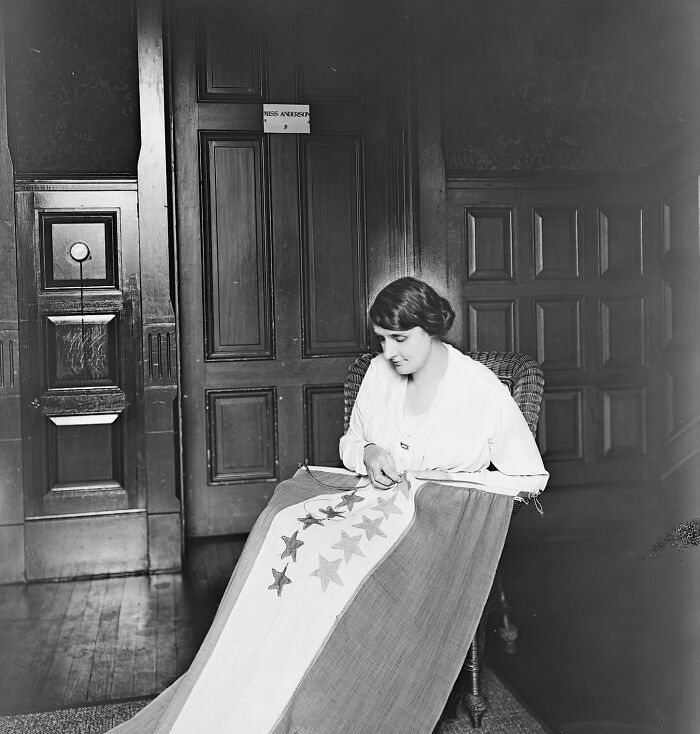 Woman sewing stars on flag representing women in the 20th century suffrage era, sitting in a wooden paneled room.