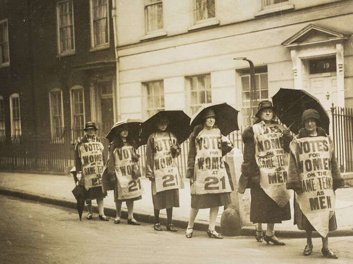 Women in the 20th century suffrage era standing in line wearing Votes for Women banners and holding umbrellas.