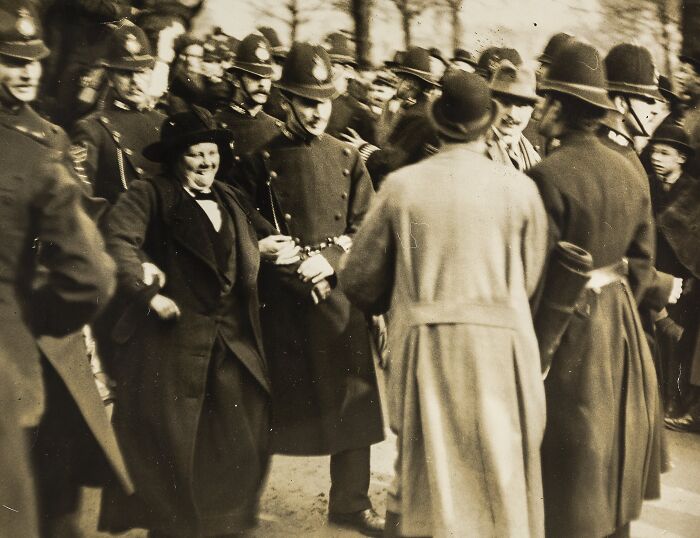 Woman being escorted by police officers during the 20th century suffrage era protest with crowd gathered around.