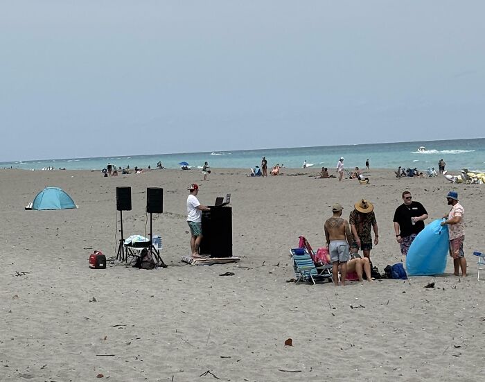 Man with DJ equipment on a busy beach, ignoring rules and etiquette as others relax nearby in chairs and tents.