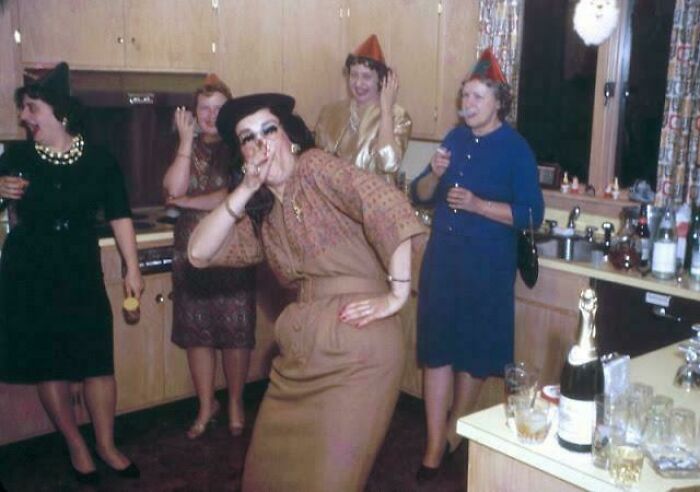 Women in vintage clothing enjoying a candid party moment in a kitchen, capturing glimpses into the past.