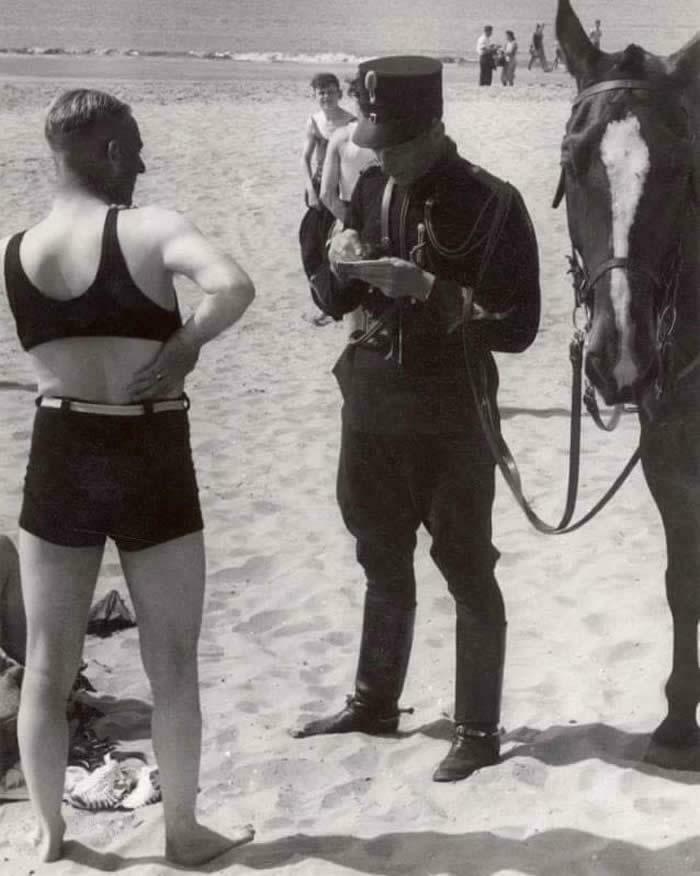 Police officer writing a ticket for a man in a swimsuit on a beach, a horse standing nearby in a candid glimpse into the past.
