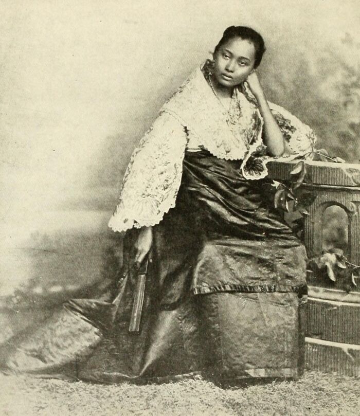 Young Filipino woman in traditional dress seated, showcasing fashion in the Philippines in the 1890s with vintage backdrop.