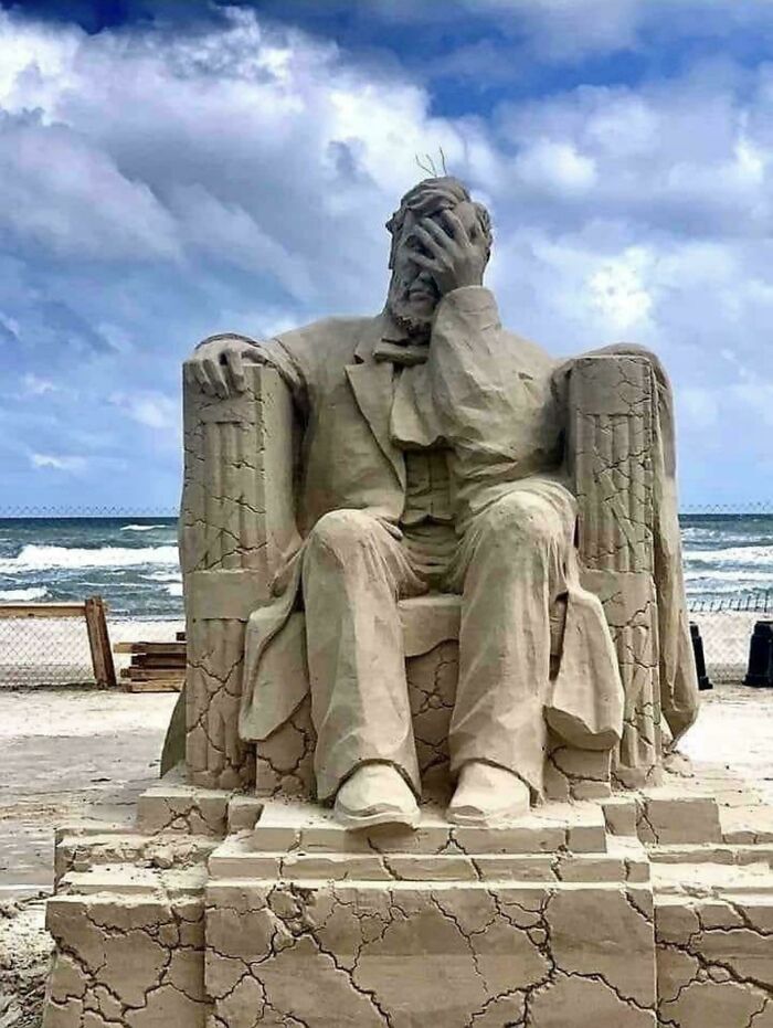 Amazing Photos sand sculpture of a seated man facepalming on a c*****d sandy pedestal by the ocean under cloudy sky